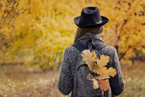 Photo of Girl in a hat with yellow leaves in a backpack is standing in the autumn forest. Back view