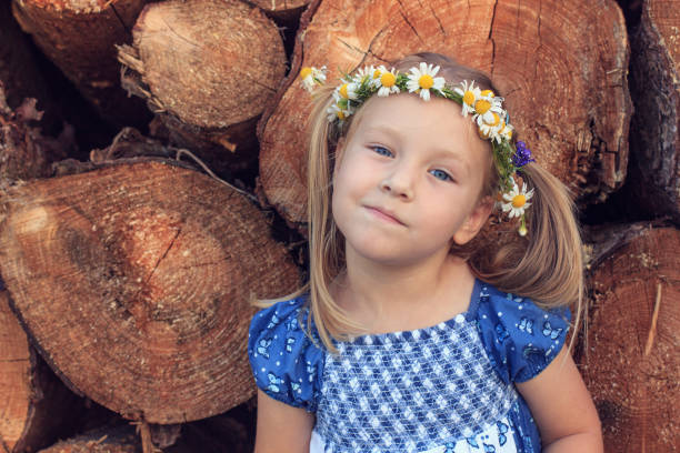 A cute 3 years old girl with chamomile wreath is standing in front of a wood stack