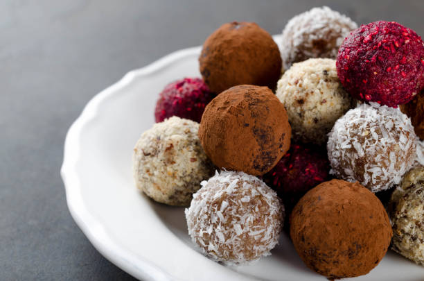 istock Homemade vegan chocolate candies with dried raspberry, grated coconut, nuts, cocoa on a white plate and dark stone background. Selective focus, copyspace 868130104