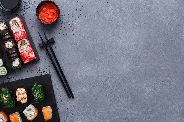 Top view set of sushi maki and rolls on rustic grey and sesame background Top view on sushi set at gray background with scattered black sesame. Slate platter with rolls, ginger and soy sauce, chopsicks on rustic surface. Japanese restaurant food and delivery, copy space restaurant table layout stock pictures, royalty-free photos & images