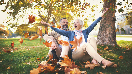 Closeup front view of early 50's couple relaxing in a park and having fun. They are sitting on the ground and throwing dry leaves.