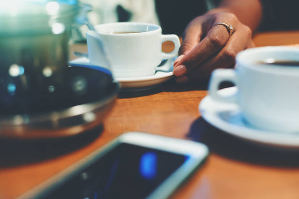 Shallow depth of field shooting of neat female hand holding plate with cup on it with delicious coffee on wooden table surrounded by another cup, glass teapot and smartphone in defocused foreground