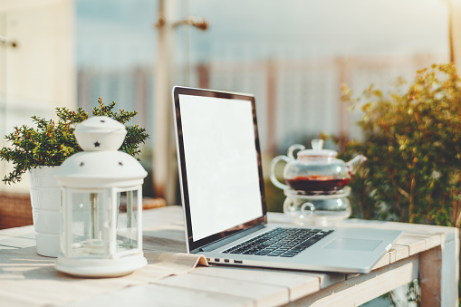 Mock-up of laptop with blank white screen standing on the wooden table outdoors in street bar; template of netbook with empty screen in cafe outdoors with teapot in defocused background on a sunny day