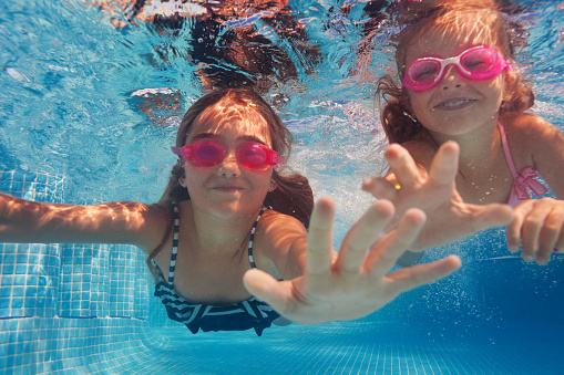 Close-up portrait of two smiling girls wearing pink goggles swimming under water of pool, pulling their hands to camera
