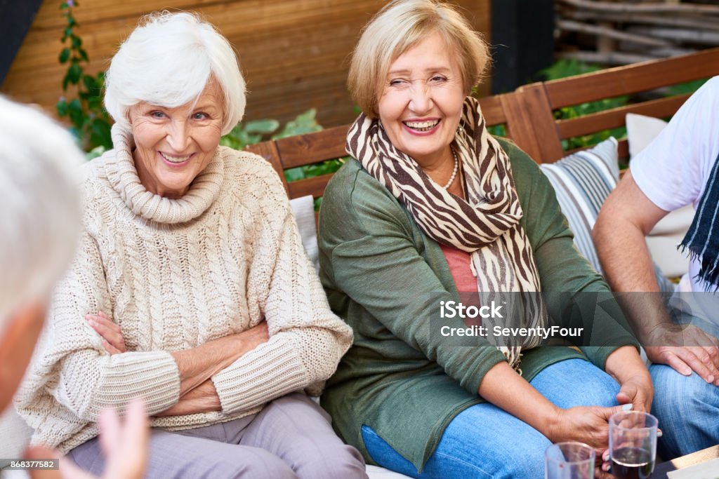 Spending Weekend with Senior Friends Pretty senior women with charming smiles listening to their male friend with interest while having gathering at cozy small patio Senior Adult Stock Photo