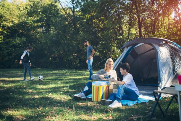 Family camping together in forest. Mother and daughter sitting in front of tent and drinking tea and coffee. Son and father playing football.