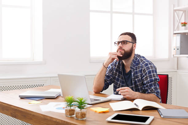 istock Pensive young businessman working with laptop in modern white office 868426464