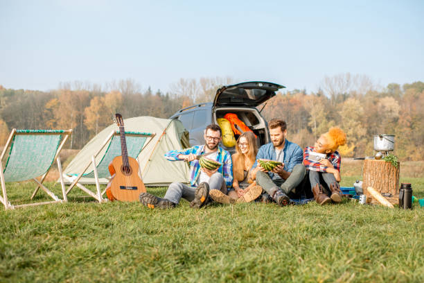 Friends during the outdoor recreation Multi ethnic group of friends having a picnic, eating watermelon, sitting in a row at the camping with tent, car and hiking equipment near the lake black man eating watermelon stock pictures, royalty-free photos & images