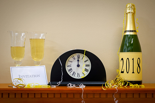 A clock showing midnight at New Year with a bottle of champagne labelled 2018, glasses and invitation surrounded by streamers.