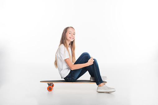 happy little female skateboarder sitting on longboard, isolated on white