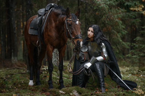 A beautiful warrior girl with a sword wearing chainmail and armor with a horse in a mysterious forest