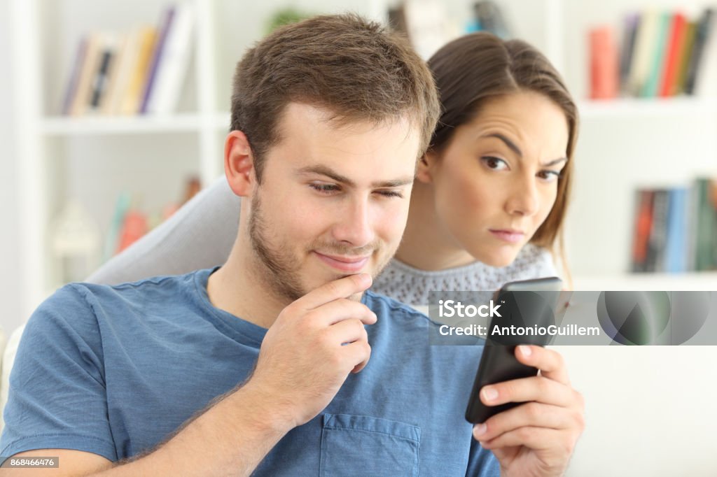 Man dating on line and girlfriend spying Cheater man dating on line with a smart phone and girlfriend is spying sitting on a sofa at home Infidelity Stock Photo