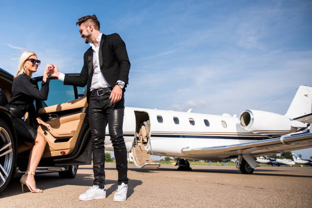Low angle view of a man helping blonde woman to get out of a car at the airport. Private jet airplane is in the background.
