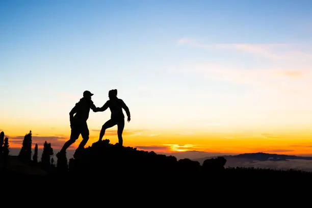 Teamwork couple with helping hand and trust. Silhouette of team help over mountains sunset. Man and woman hikers or climbers reaching goals in inspirational landscape on Tenerife Canary Islands.