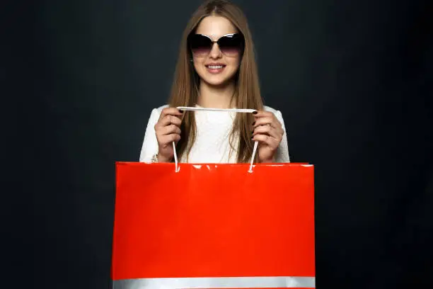 Stylish girl in sunglasses and a white dress, holds red package for shopping, isolated against a dark background. Template. Mock-up