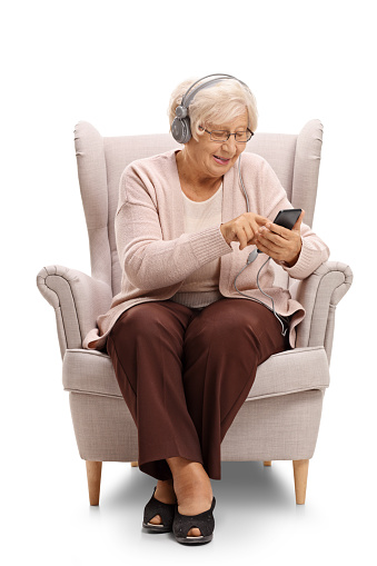 Elderly woman sitting in an armchair and listening to music on a phone isolated on white background