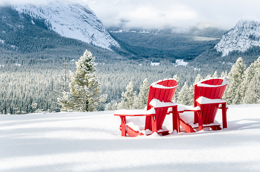 Photo of Two Snow Covered Red Adirondack Chairs Facing a Frozen Wooded Valley on a Snowy Winter Day. Banff National Park, AB, Canada.