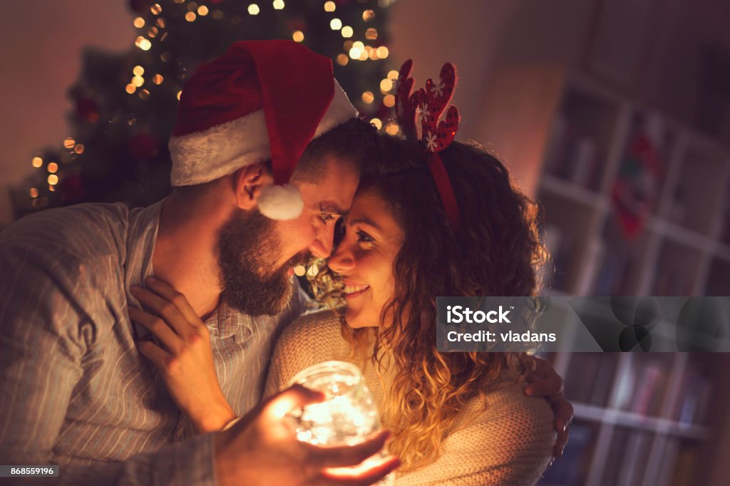 Christmas romance Beautiful couple in love sitting on the living room floor next to a Christmas tree, holding a jar with Christmas lights Christmas Stock Photo