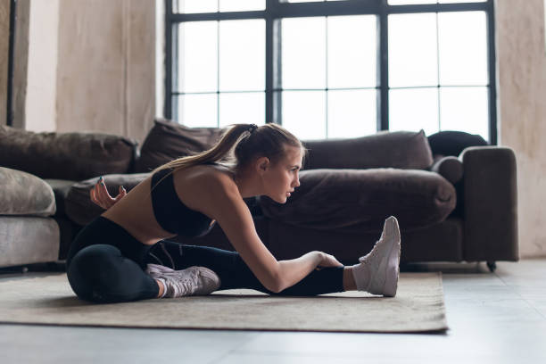 istock Slim sportswoman in sportswear warming-up muscles before workout doing stretching exercise sitting on floor at home 868604288