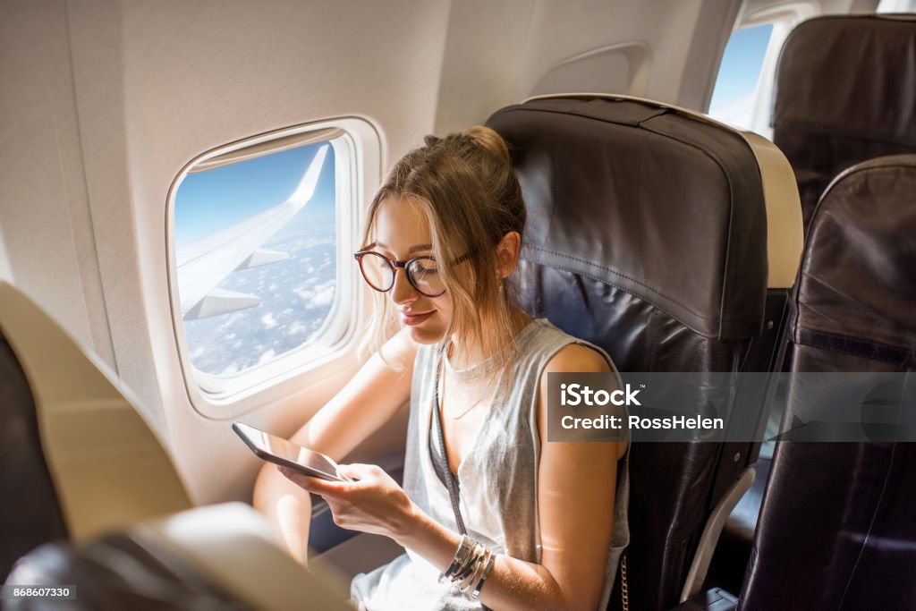 Woman in the airplane Young woman sitting with phone on the aircraft seat near the window during the flight in the airplane Airplane Stock Photo