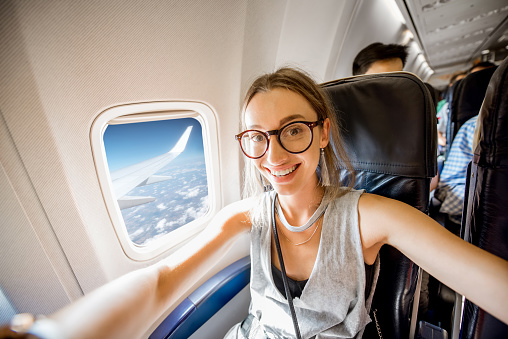 Young happy woman making selfie photo sitting on the aircraft seat near the window during the flight in the airplane