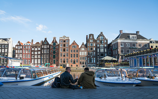 AMSTERDAM, HOLLAND - AUGUST 19, 2017; Two men sitting on shady side of canal across from architectural facades of a range of styles for which the city is known on other side of canal across from Damrak