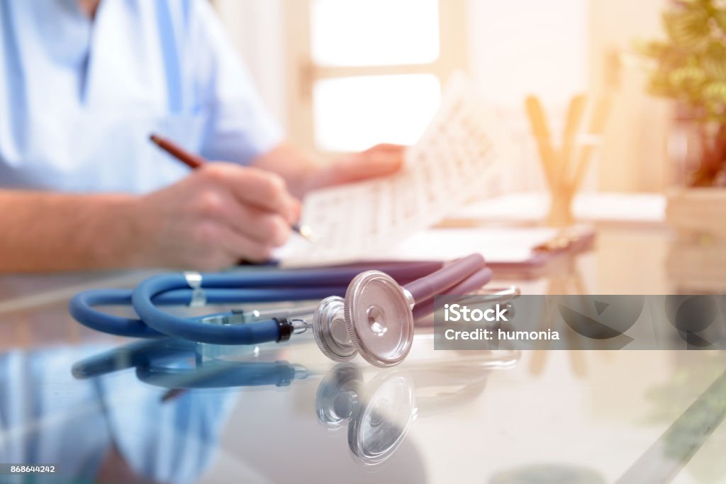 Close up of stethoscope and doctor Close up of stethoscope and doctor sitting in a background filling in documentation Doctor Stock Photo