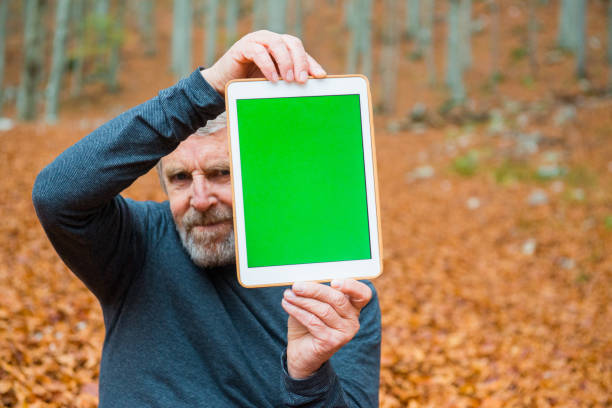 Senior Man Holding Digital Tablet with Green Screen and Partly Hiding his Face, Autumnal Beech Forest, Europe Senior man holding digital tablet with green screen in front and partly hiding his face in autumnal beech forest, Europe. person holding frame over face stock pictures, royalty-free photos & images
