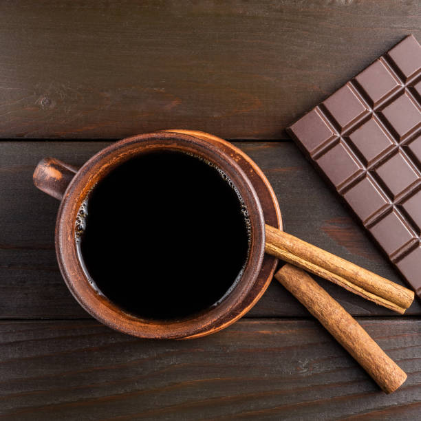 Hot black coffee in brown clay cup, dark chocolate bar and cinnamon stick on wooden background, top view
