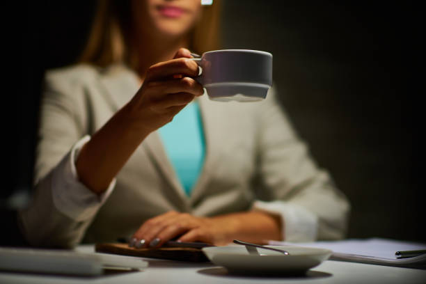 Close-up of business lady holding coffee cup while drinking coffee at work in dark office