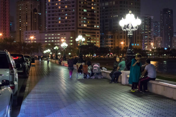 A well-maintained promenade runs along a river, highlighted by a contemporary trashcan. In the background, buildings and a small house complement the tranquil waters under a clear sky.