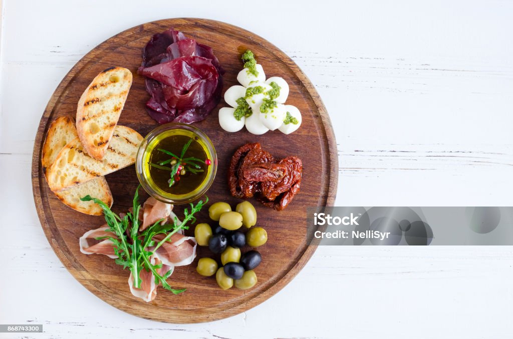 Meat and cheese plate antipasti snack Meat and cheese plate antipasti snack with Prosciutto ham, arugula, bresaola, Mozzarella balls with pesto sauce, sun-dried tomatoes, olives and toasts with spices olive oil on wooden board. Appetizer Stock Photo