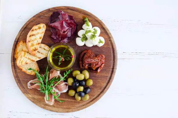 Meat and cheese plate antipasti snack with Prosciutto ham, arugula, bresaola, Mozzarella balls with pesto sauce, sun-dried tomatoes, olives and toasts with spices olive oil on wooden board.