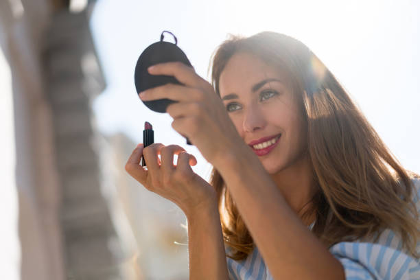 Portrait of a teenage girl applying red lipstick outdoors in a woodland location. She is looking at the mirror on her smart phone to make sure she applies the lipstick evenly. The girl has brown hair with red auburn highlights. Focus is on the girl while the lush green of the forest is defocused beyond.