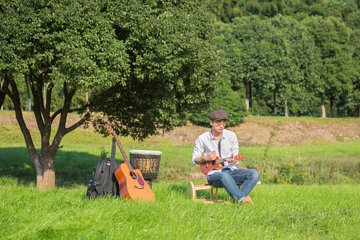 young man playing small guitar on lawn,with djembe drum and acoutic guitar on side.