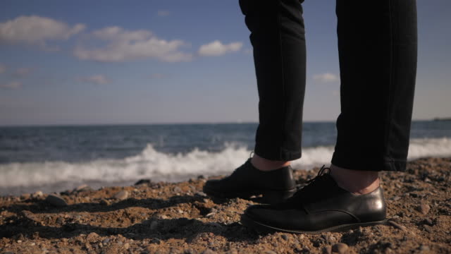 Woman legs standing on sandy beach near sea with waves. Female in leather shoes on shore. Close up of girls feet. Beautiful legs on border sea. Slow motion