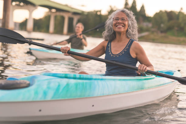 An ethnic senior woman smiles while kayaking with her husband A mixed race senior couple is kayaking down a river together. The image's focus is on the elderly woman smiling in the foreground. She is Pacific Islander and has graying hair. Her husband is seen in a kayak in the background. In the distance behind the couple you can see the shoreline and a bridge. The happy couple are wearing casual clothing. It is warm outside. health lifestyle stock pictures, royalty-free photos & images