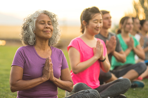 A senior woman of Pacific Islander descent meditates during a group yoga class. She has a slight smile and is seated in the prayer pose. The multi-ethnic group is outdoors. Men and woman of different ages are taking the class. The class participants are seated in a line and they are all doing the prayer pose.