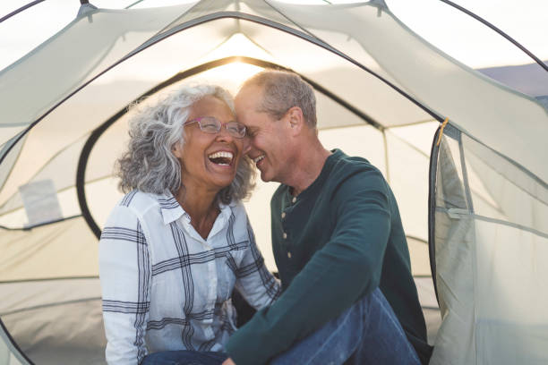 Retirement doesn't get much sweeter than this A mixed race senior couple snuggle and flirt. They are camping and are seated in their tent. The door to their tent is unzipped. Light from the setting sun shines through the back of the tent. The couple is laughing. The husband has his eyes closed and is resting his forehead on his wife's temple. The wife is looking off into the distance while she laughs. The couple is dressed in casual clothing and the woman is wearing glasses. baby boomer stock pictures, royalty-free photos & images
