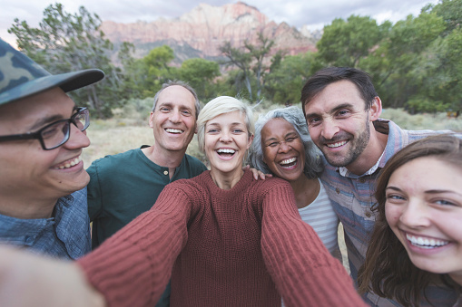 A mixed race senior couple pose with their adult children and friends for a group photo outdoors. A young woman at the center of the frame is holding out the camera and is taking a selfie of the group. Everyone is smiling and laughing. They are standing in a field embracing one another. A forest and mountains are in the background. It is near sunset. The group is on vacation together.