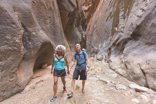 An ethnic senior couple explore a slot canyon together. They are hiking down a river and are pictured wading. The couple is wearing casual clothing and swimwear. The woman leads the way as she holds her husband's hand. She is smiling and looking ahead. The man is looking up and admiring the unique landscape.