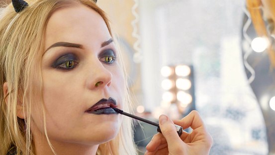 Process applying halloween makeup on face the young beautiful blonde woman contact lenses as cat eyes. Artist applying lipstick.