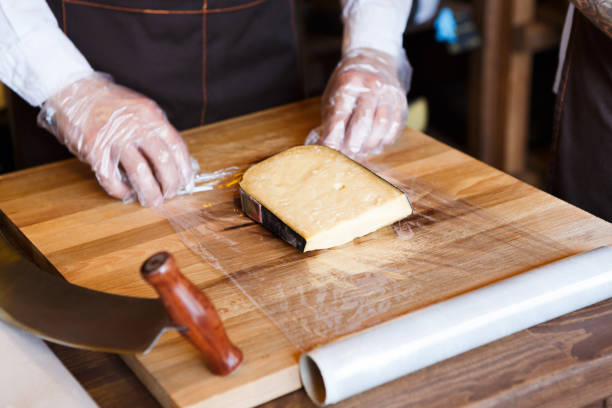Shop assistant wrapping a piece of cheese. Packing order at grocery shop in cling film on wooden table, background