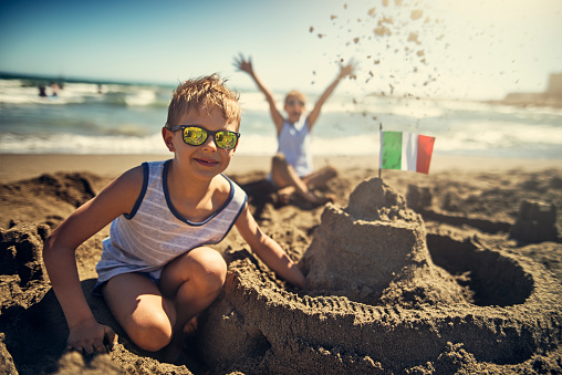 Happy boys are building sandcastle on a beach on a sunny summer day. They sticked a italian flag in the sandcastle.