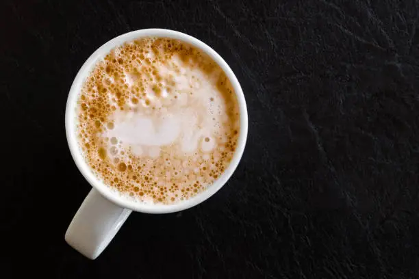 Milky frothy coffee in white mug isolated on black leather from above.