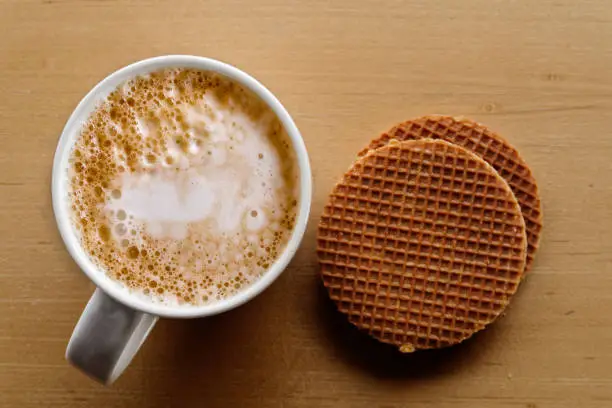 Milky frothy coffee in white mug next to a couple of round waffle biscuits isolated on light wood from above.