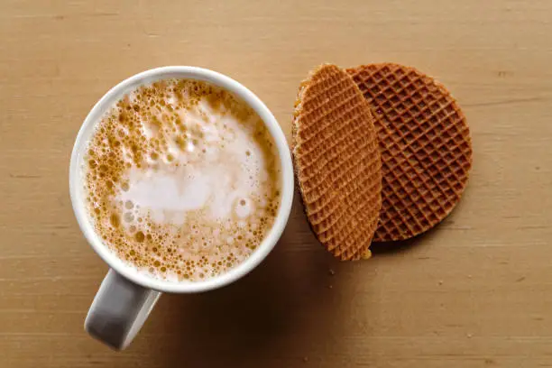 Milky frothy coffee in white mug next to a couple of round waffle biscuits isolated on light wood from above.
