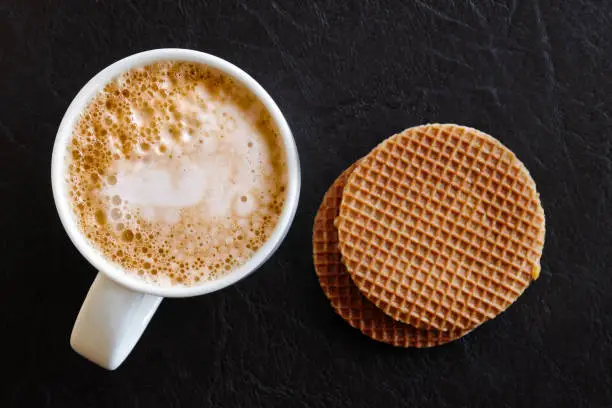 Milky frothy coffee in white mug next to a couple of round waffle biscuits isolated on black leather from above.