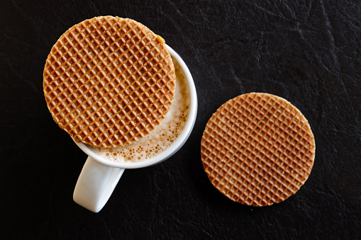 White mug with milky frothy coffee and a round waffle biscuit on top next to another waffle biscuit isolated on black leather from above.