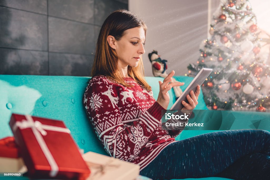 Women using tablet during christmas Women sitting on the blue sofa by the christmas tree and using tablet Adult Stock Photo